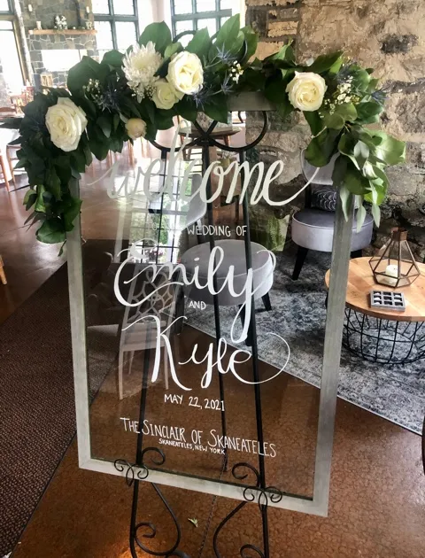 A sign decorate with white flowers that says "Welcome Emily and Kyle"